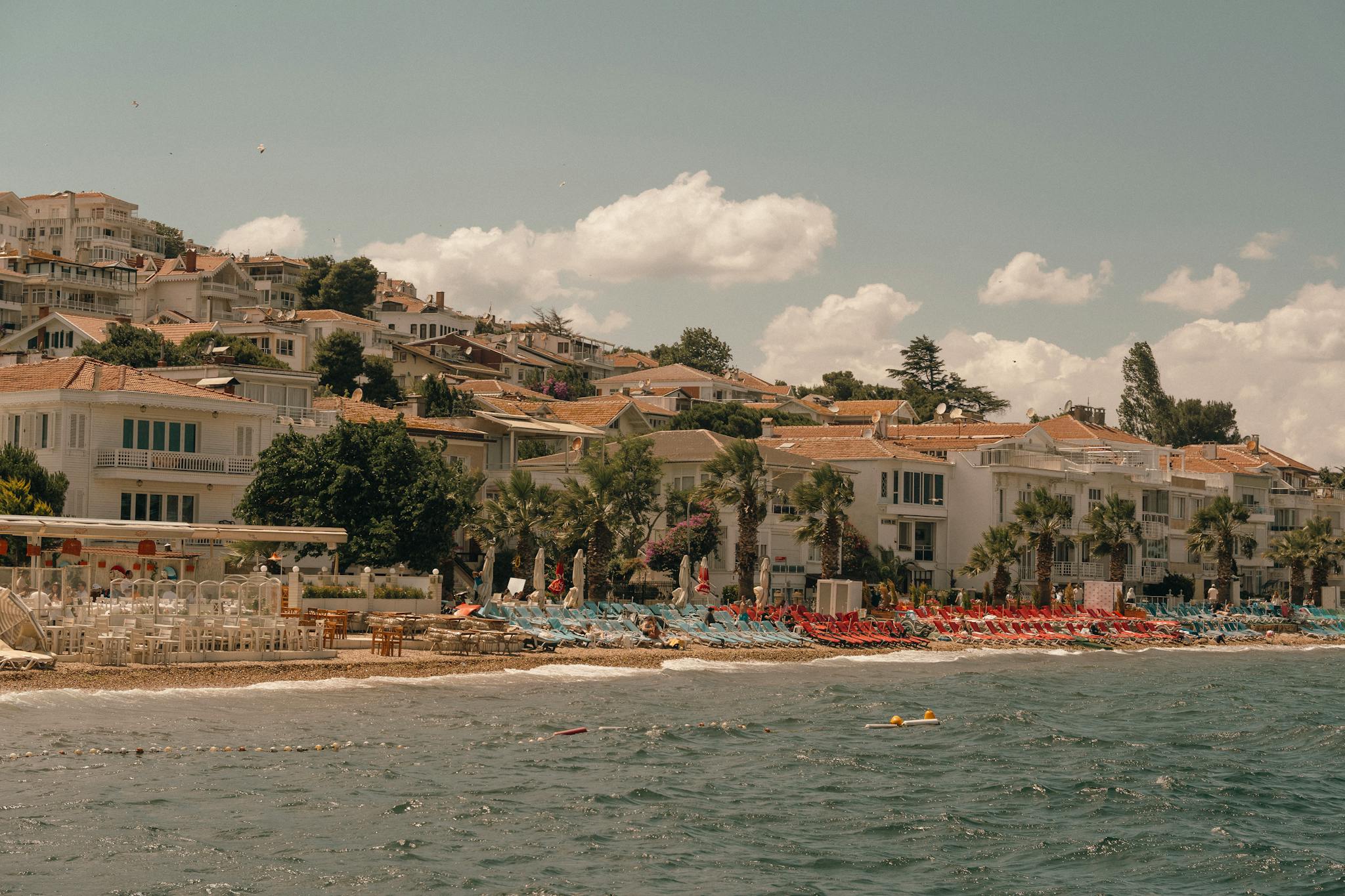 Charming coastal scene with Mediterranean villas, clear skies, and vibrant beach chairs along the shoreline.